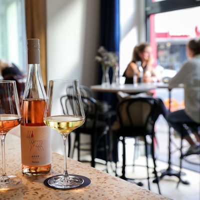 03.07.2023, Köln: Die Weinbar Bedrouille: Auf der Theke steht eine Flasche Wein mit Gläsern, im Hintergund sitzen Menschen an Tischen.