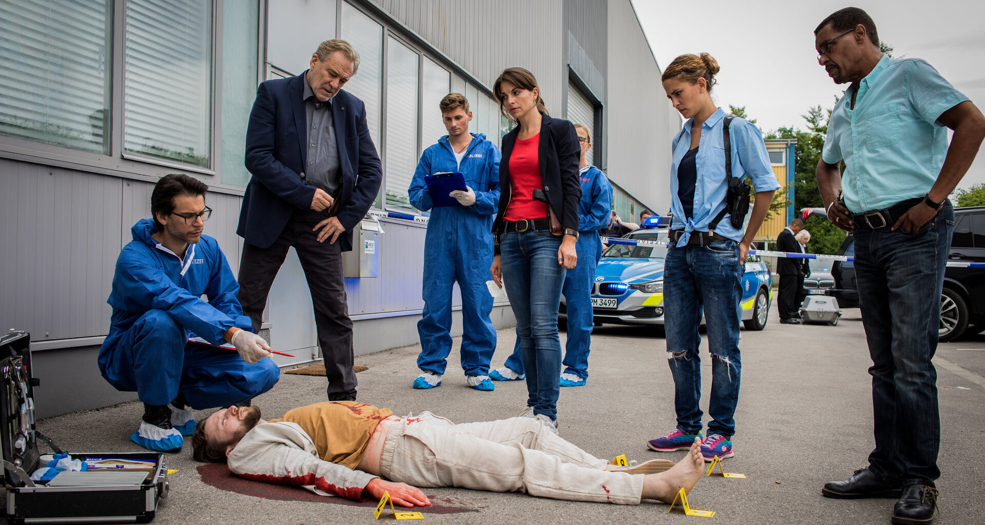 "SOKO München - 24 Stunden": Am Tatort auf einem grauen Industriegelände liegt eine Leiche auf dem Boden. Um diese herum steht das SOKO-Team bestehend aus Arthur Bauer (Gerd Silberbauer), Katharina Hahn (Bianca Hein), Toni Bischoff (Amanda da Gloria) und Franz Ainfachnur (Christofer v. Beau) und schauen sich die Leiche näher an. Dr. Weissenböck (Florian Odendahl) sitzt in der Hocke am Kopf der Leiche, er hält einen Stift in der Hand. Im Hintergrund sind zwei Mitarbeiter der Spusi (Komparsen) und ein Polizeiauto zu sehen.