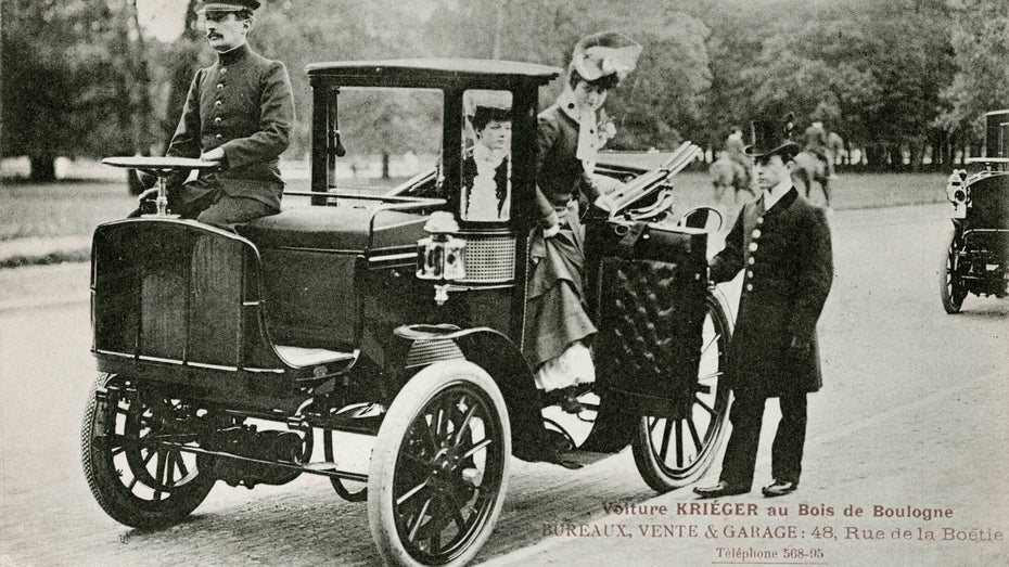 Schwarz-Weiß: Zwei Frauen werden von einem Chauffeur in einem Krieger-Elektroauto gefahren