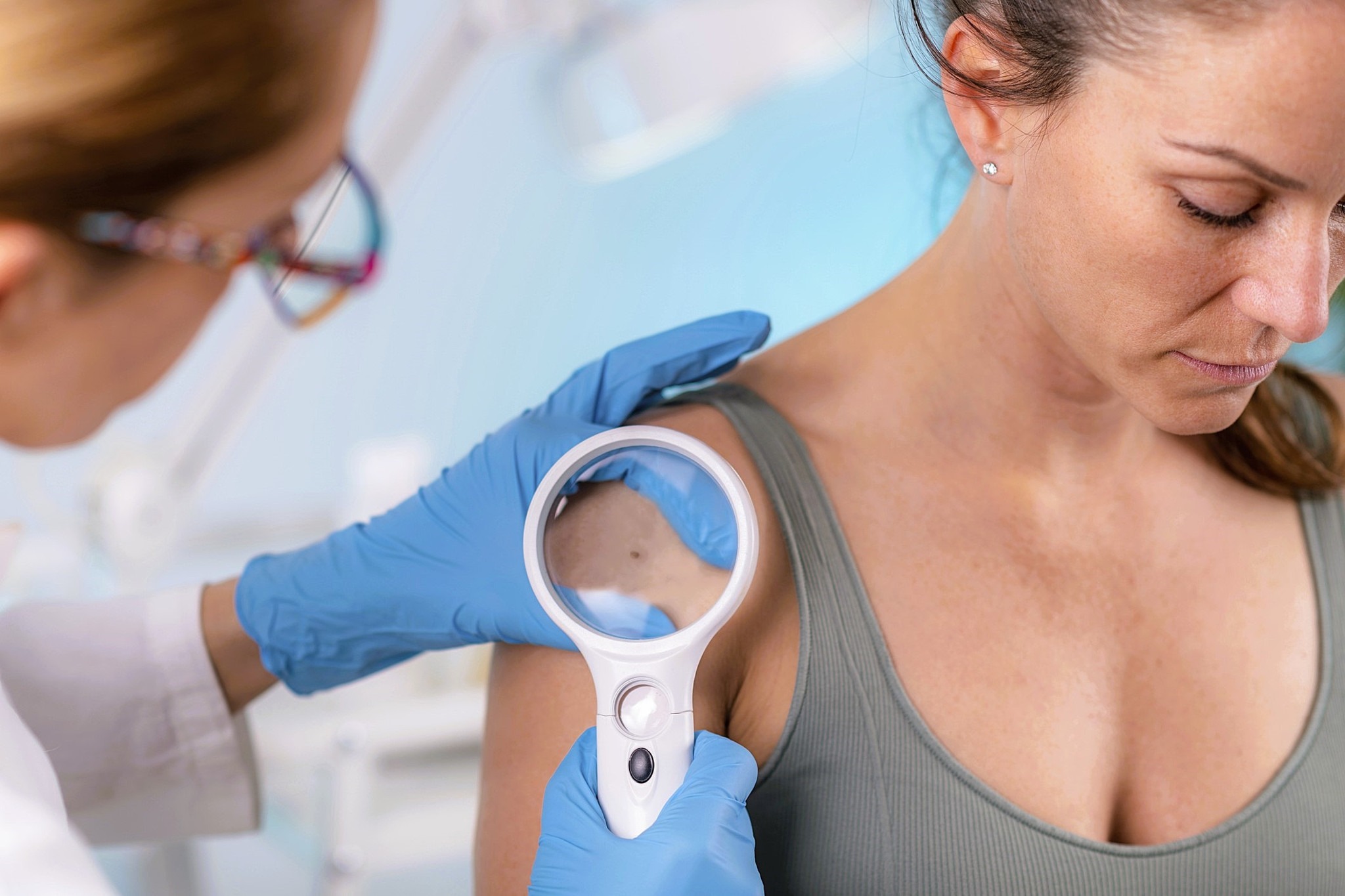 Eine Dermatologin hält ein Dermatoskop in der rechten Hand und untersucht die Haut einer Patientin am Oberarm.