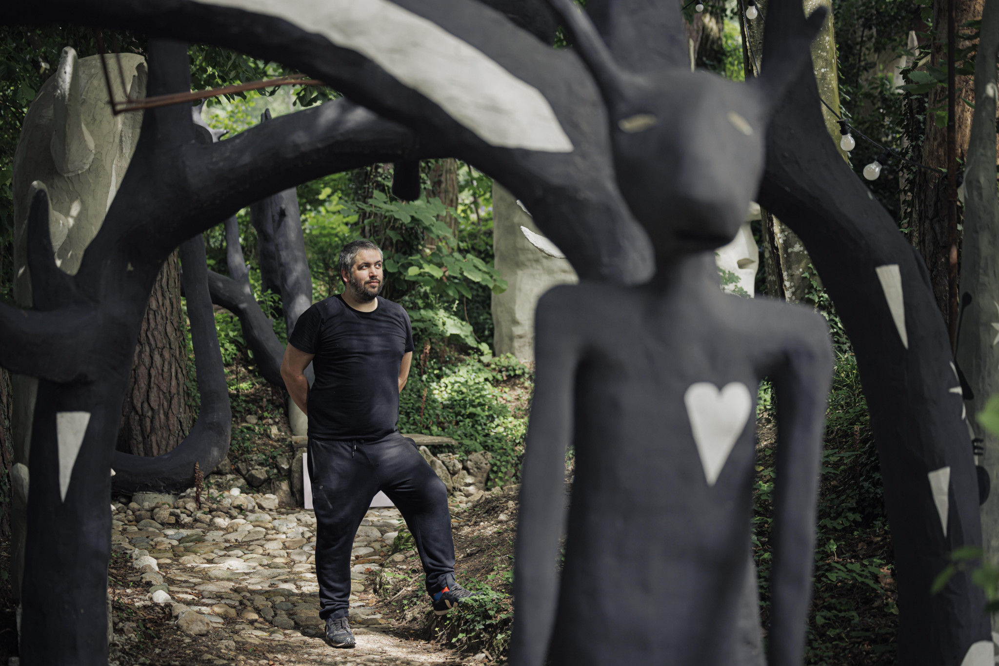 Ein Mann steht in einem Wald, umgeben von schwarzen Skulpturen mit Herzmotiven.