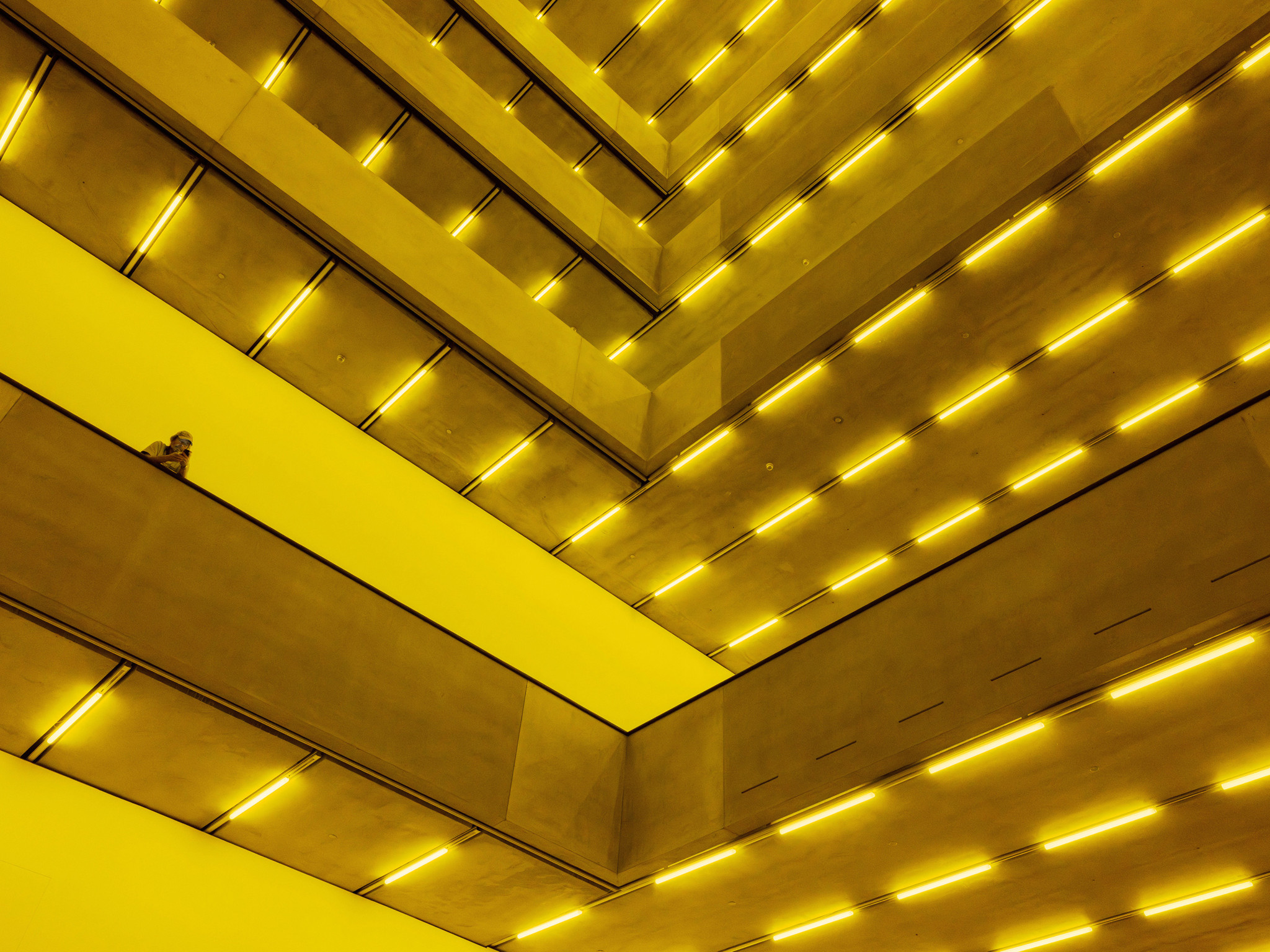 Modernes Gebäude mit leuchtend gelben Beleuchtungselementen an den Decken. Mehrere Ebenen und Perspektiven schaffen ein futuristisches Design.