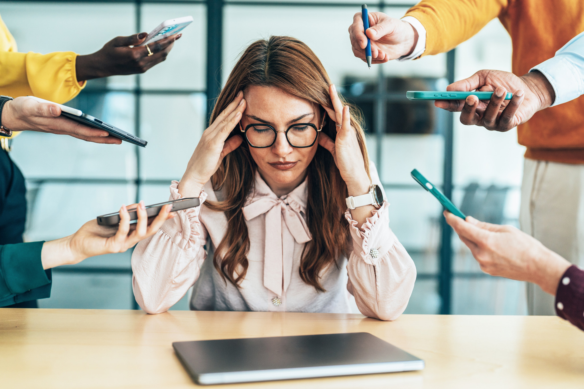 Eine Frau mit langem braunen Haar sitzt in einem Büro an einem Schreibtisch, hält beide Hände an ihre Schläfen und schaut bedrückt nach unten. Vor ihr liegt ein Laptop. Rechts und links sind je drei Hände mit Smartphones und Stiften gegen die Frau gerichtet. 