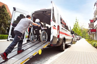 Johanniter Fahrdienstfahrer schiebt ältere Frau im Rollstuhl in Transporter, Unterstützung im Alltag