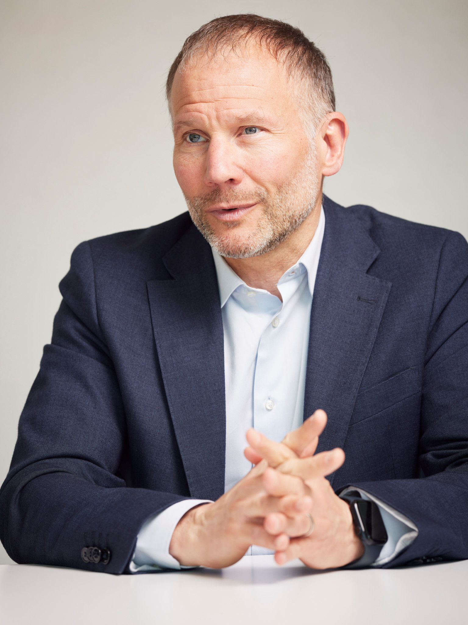 Ein Mann mit blauen Augen, lichtem Haar und Dreitagebart sitzt an einem Tisch. Er trägt ein hellblaues Hemd und ein blaues Sakko. 