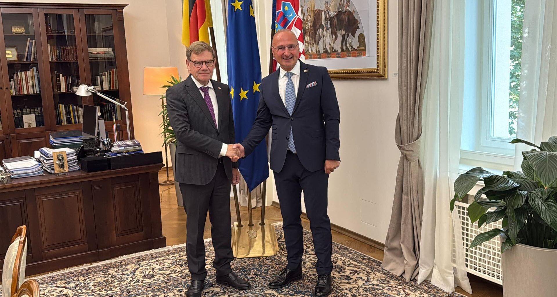 Die beiden stehen im Arbeitszimmer des kroatischen Ministers, schütteln sich die Hände und schauen lächelnd in die Kamera. Hinter ihnen die Flaggen ihrer Länder sowie der EU, außerdem ein Schreibtisch.