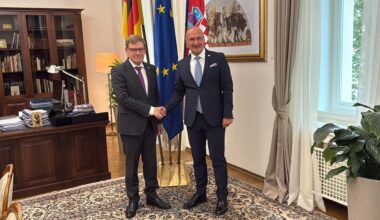 Die beiden stehen im Arbeitszimmer des kroatischen Ministers, schütteln sich die Hände und schauen lächelnd in die Kamera. Hinter ihnen die Flaggen ihrer Länder sowie der EU, außerdem ein Schreibtisch.