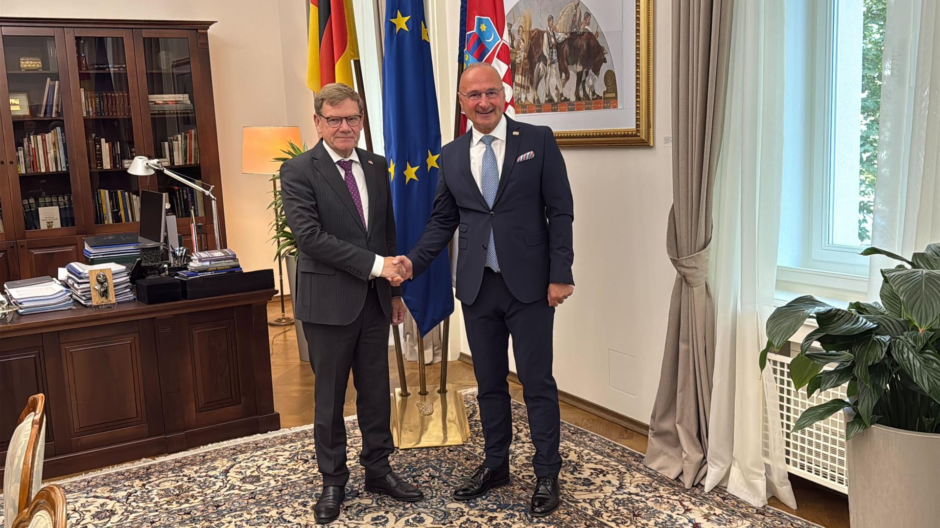 Die beiden stehen im Arbeitszimmer des kroatischen Ministers, schütteln sich die Hände und schauen lächelnd in die Kamera. Hinter ihnen die Flaggen ihrer Länder sowie der EU, außerdem ein Schreibtisch.