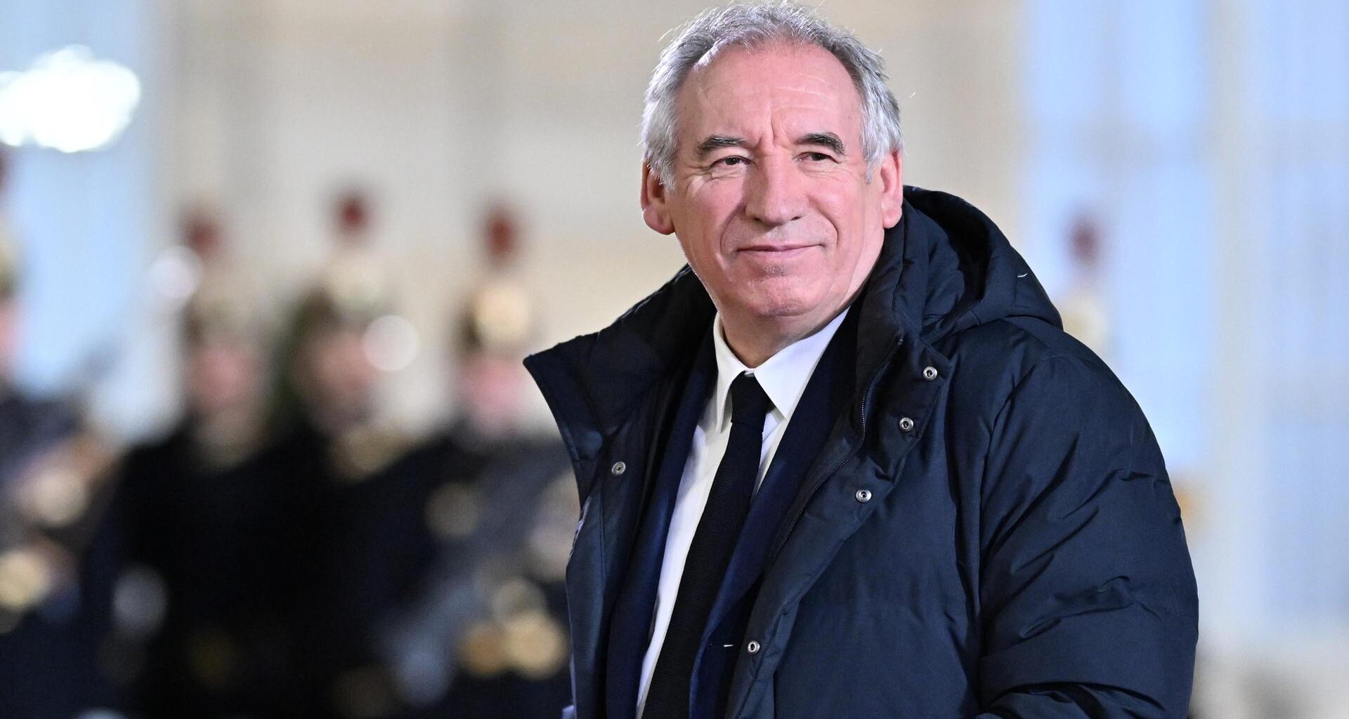 Bayrou trägt einen langen Daunenanorak und guckt in die Kamera. Dahinter unscharf Mitglieder einer Musikkapelle.
