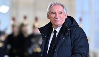 Bayrou trägt einen langen Daunenanorak und guckt in die Kamera. Dahinter unscharf Mitglieder einer Musikkapelle.