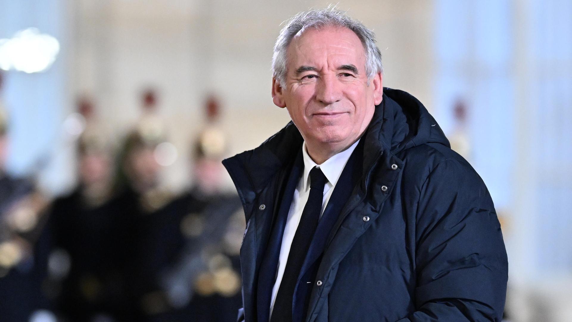 Bayrou trägt einen langen Daunenanorak und guckt in die Kamera. Dahinter unscharf Mitglieder einer Musikkapelle.