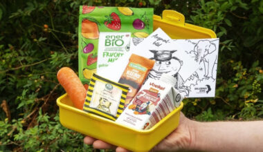 Biobrotbox 2025 mit gesunden Snacks, Karotte, Bio-Fruchtsnack, Müsliriegel, Getränk und Ausmalbild in gelber Brotdose