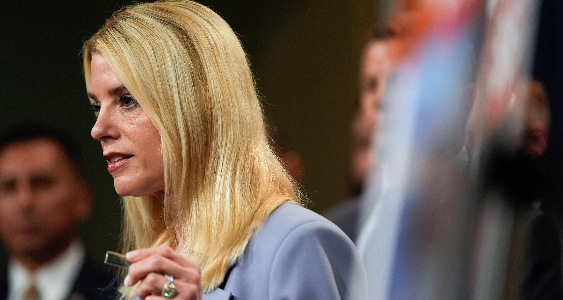 Man sieht die blonde langhaarige Bondi im hellen Jackett von der Seite. Sie sagt etwas und hebt die linke Hand.  Dahinter unscharf weitere Personen.