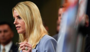 Man sieht die blonde langhaarige Bondi im hellen Jackett von der Seite. Sie sagt etwas und hebt die linke Hand.  Dahinter unscharf weitere Personen.