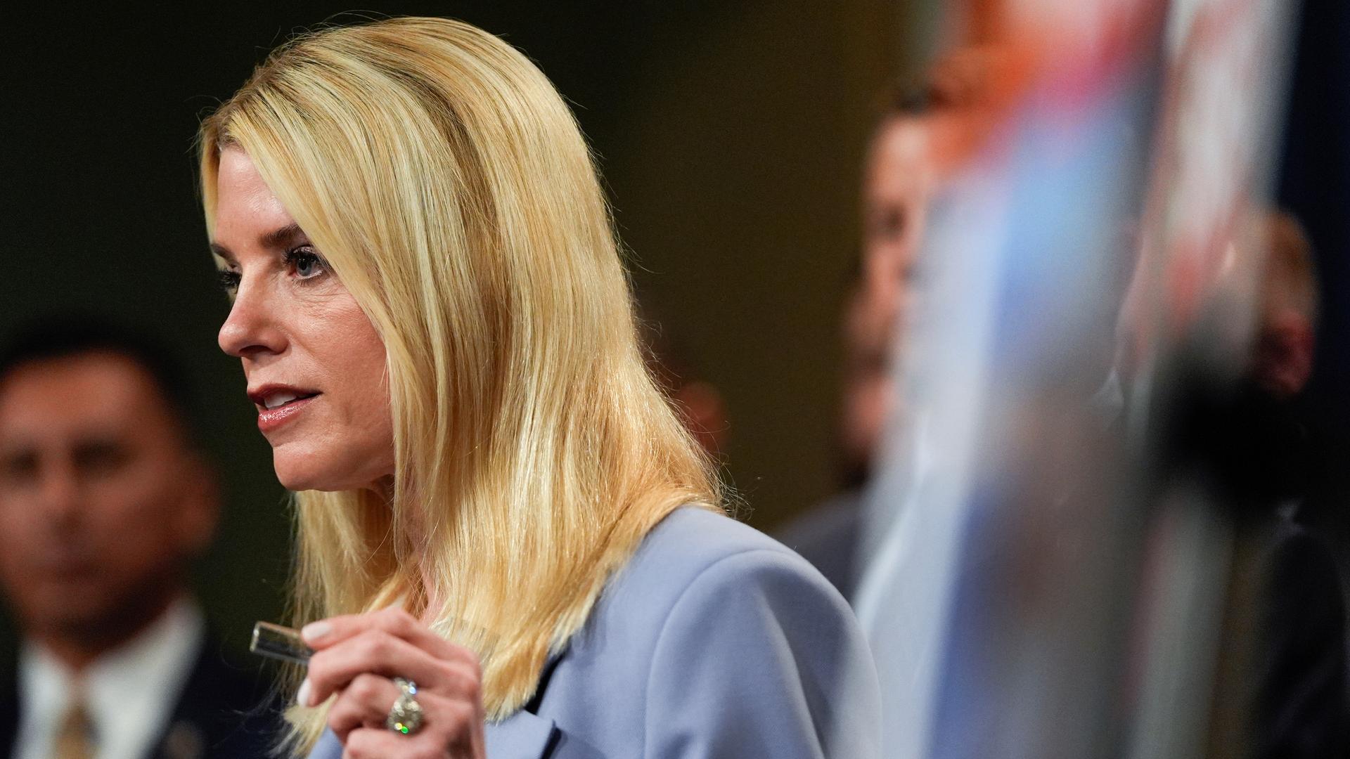 Man sieht die blonde langhaarige Bondi im hellen Jackett von der Seite. Sie sagt etwas und hebt die linke Hand.  Dahinter unscharf weitere Personen.