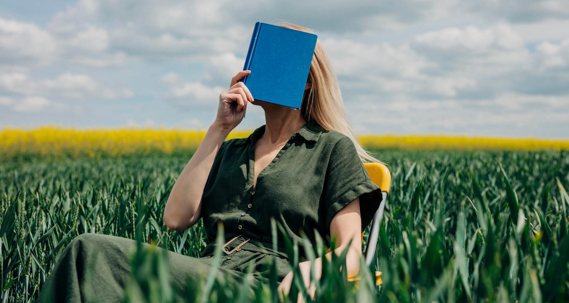 Eine Frau sitzt mit einem blauen Buch vor dem Gesicht in einem Feld auf einem Stuhl.