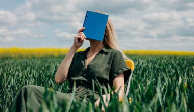 Eine Frau sitzt mit einem blauen Buch vor dem Gesicht in einem Feld auf einem Stuhl.