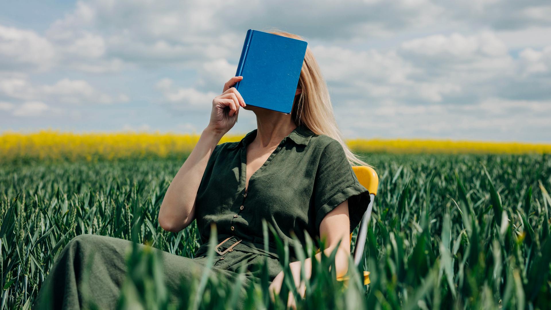 Eine Frau sitzt mit einem blauen Buch vor dem Gesicht in einem Feld auf einem Stuhl.