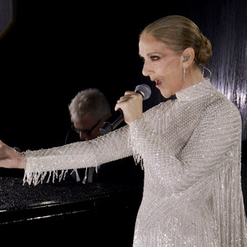 Céline Dion hält einen Arm ausgestreckt während sie vor einem Pianisten auf der ersten Etage des Eiffelturms singt. Sie trägt ein weißes Perlenkleid von Dior.