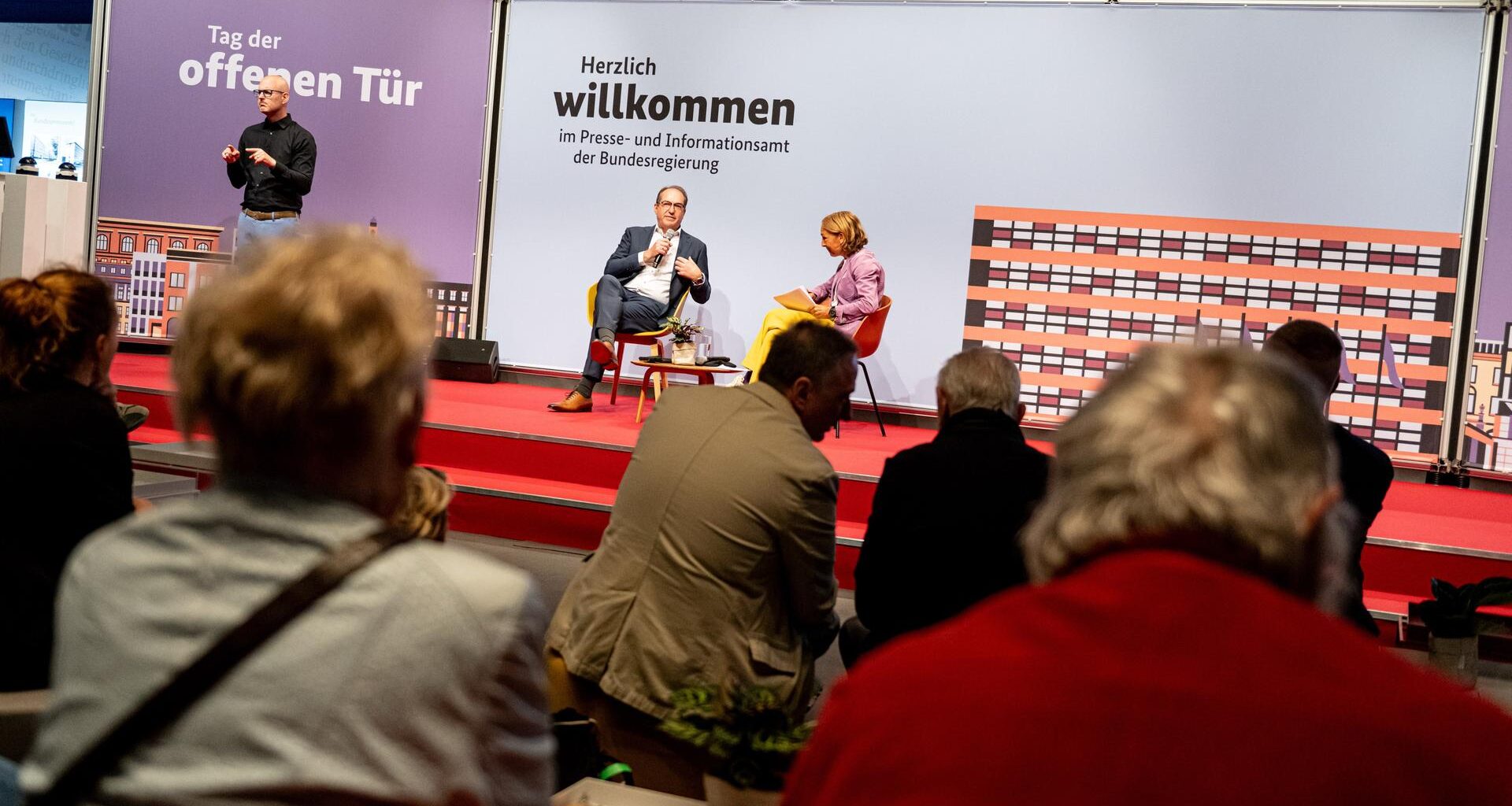Innenminister Dobrindt sitzt im Bildhintergrund auf einer Bühne mit einem Mikrofon in der Hand. Neben ihm sitzt eine Frau, die ihn befragt. Eine Gruppe von Menschen hört zu. Auf einer Stellwand steht "Tag der offenen Tür" und "Herzlich Willkommen".