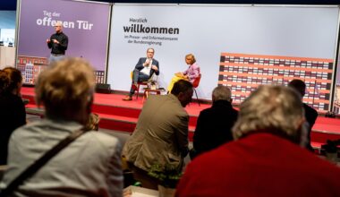 Innenminister Dobrindt sitzt im Bildhintergrund auf einer Bühne mit einem Mikrofon in der Hand. Neben ihm sitzt eine Frau, die ihn befragt. Eine Gruppe von Menschen hört zu. Auf einer Stellwand steht "Tag der offenen Tür" und "Herzlich Willkommen".