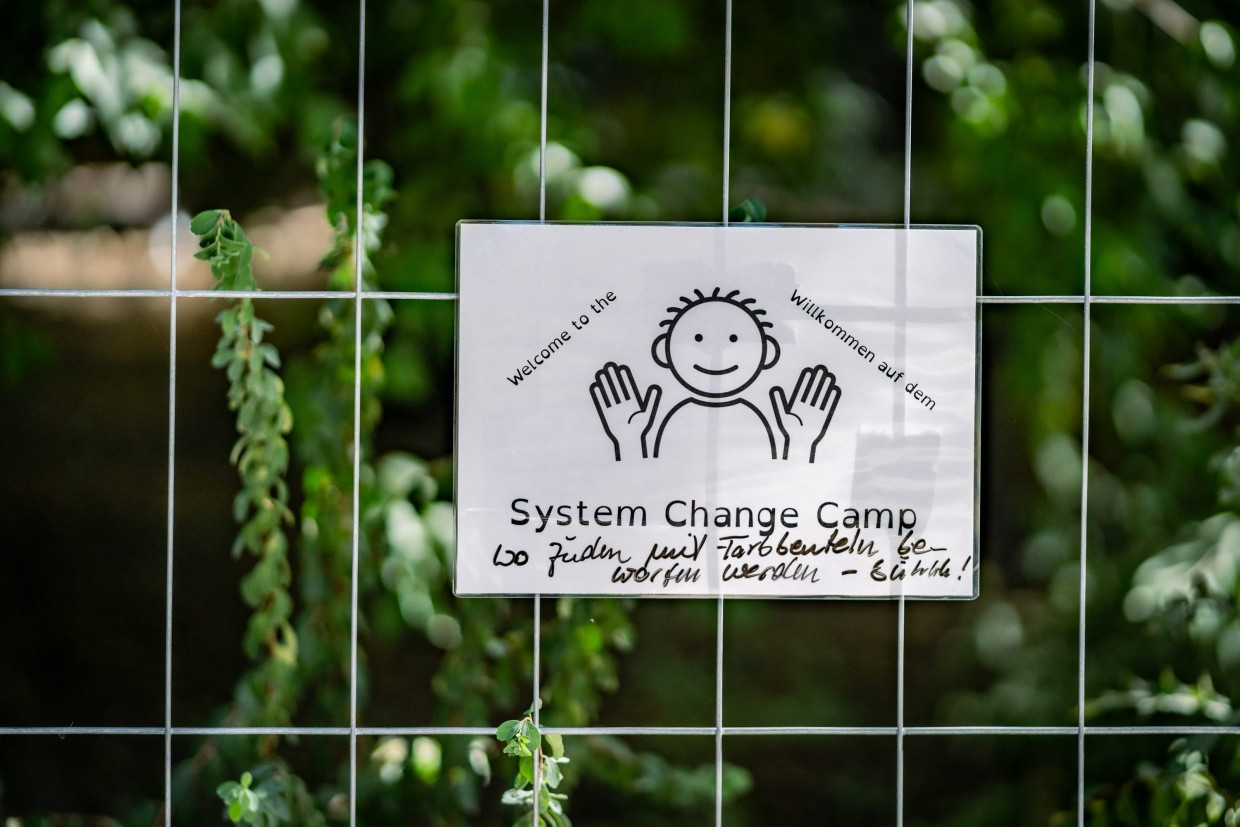 Ein kleines Plakat mit der Aufschrift „Willkommen auf dem System Change Camp” und der handschriftlichen Ergänzung „Wo Juden mit Farbbeuteln beworfen werden”.
