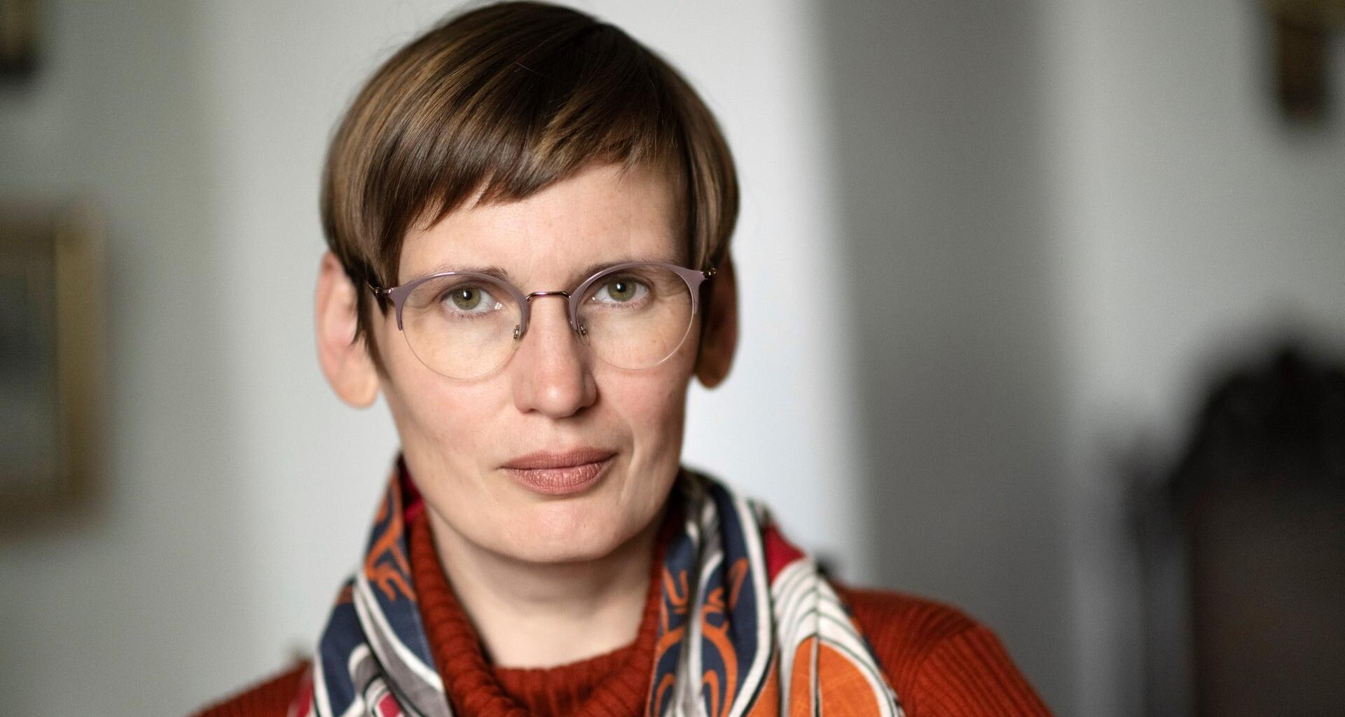 Eine Frau mit dunkelblonder Kurzhaarfrisur und runder Brille schaut nachdenklich in die Kamera. Sie trägt einen orangefarbenen Rollkragenpullover und einen bunt gemusterten Schal.