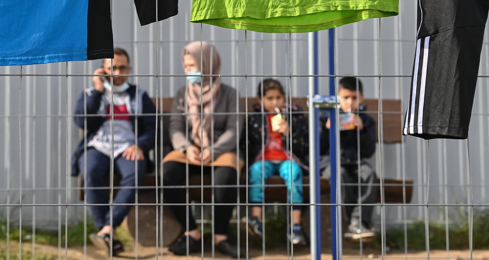 Eine Familie mit zwei Kindern ist unscharf hinter einem Zaun zu sehen. Sie sitzen nebeneinander auf einer Bank vor einer Contanierwand.
