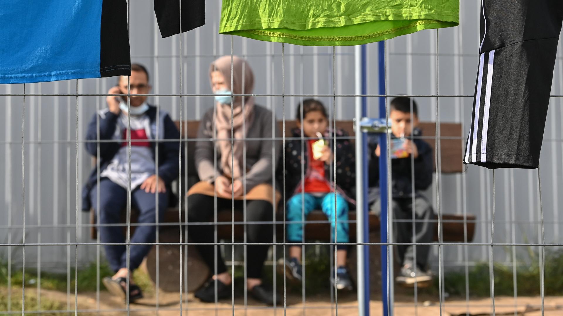Eine Familie mit zwei Kindern ist unscharf hinter einem Zaun zu sehen. Sie sitzen nebeneinander auf einer Bank vor einer Contanierwand.