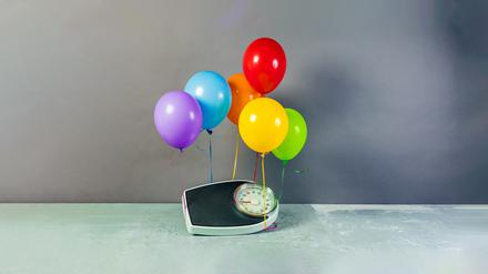 Eine Personenwaage wird von bunten Heliumluftballons angehoben.