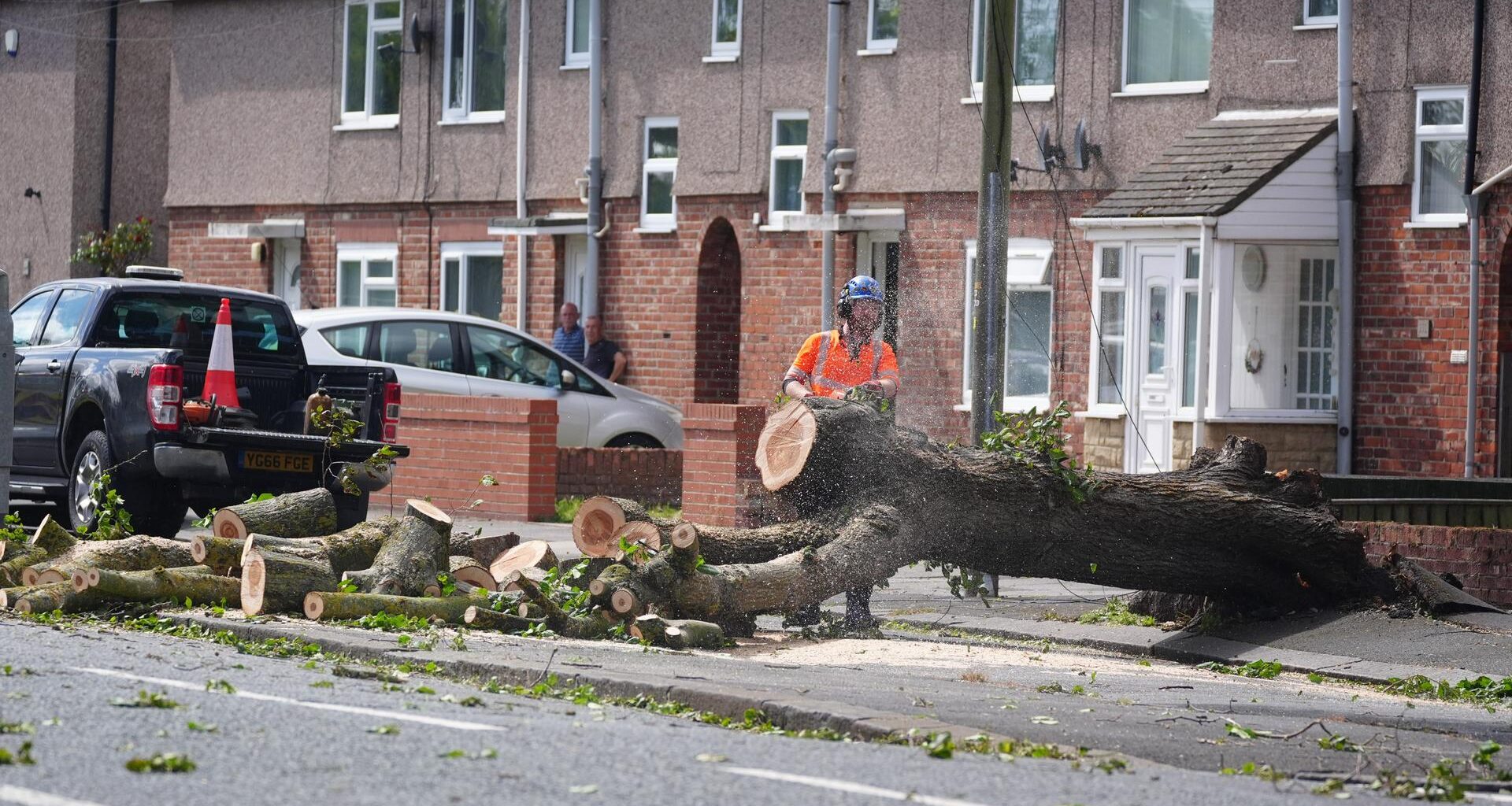 Ein Feuerwehrmann zerschneidet einen umgestürzten Baum in einer Straße mit Reihenhäusern.