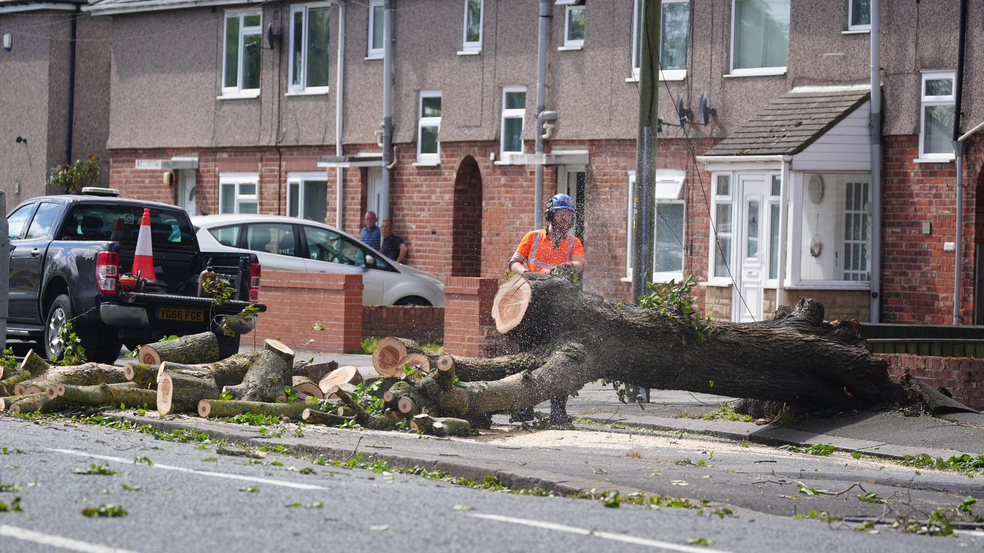 Ein Feuerwehrmann zerschneidet einen umgestürzten Baum in einer Straße mit Reihenhäusern.