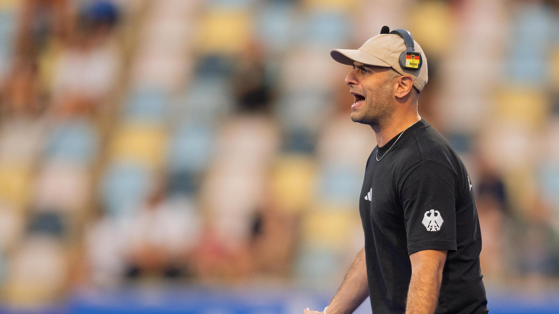 Hockey-Bundestrainer Henning ruft etwas vom Spielfeld-Rand. Er hat ein schwarzes Trikot an und eine beige Kappe auf. Und trägt darüber einen Kopfhörer mit Deutschland-Farben. Henning sieht frustriert aus. 