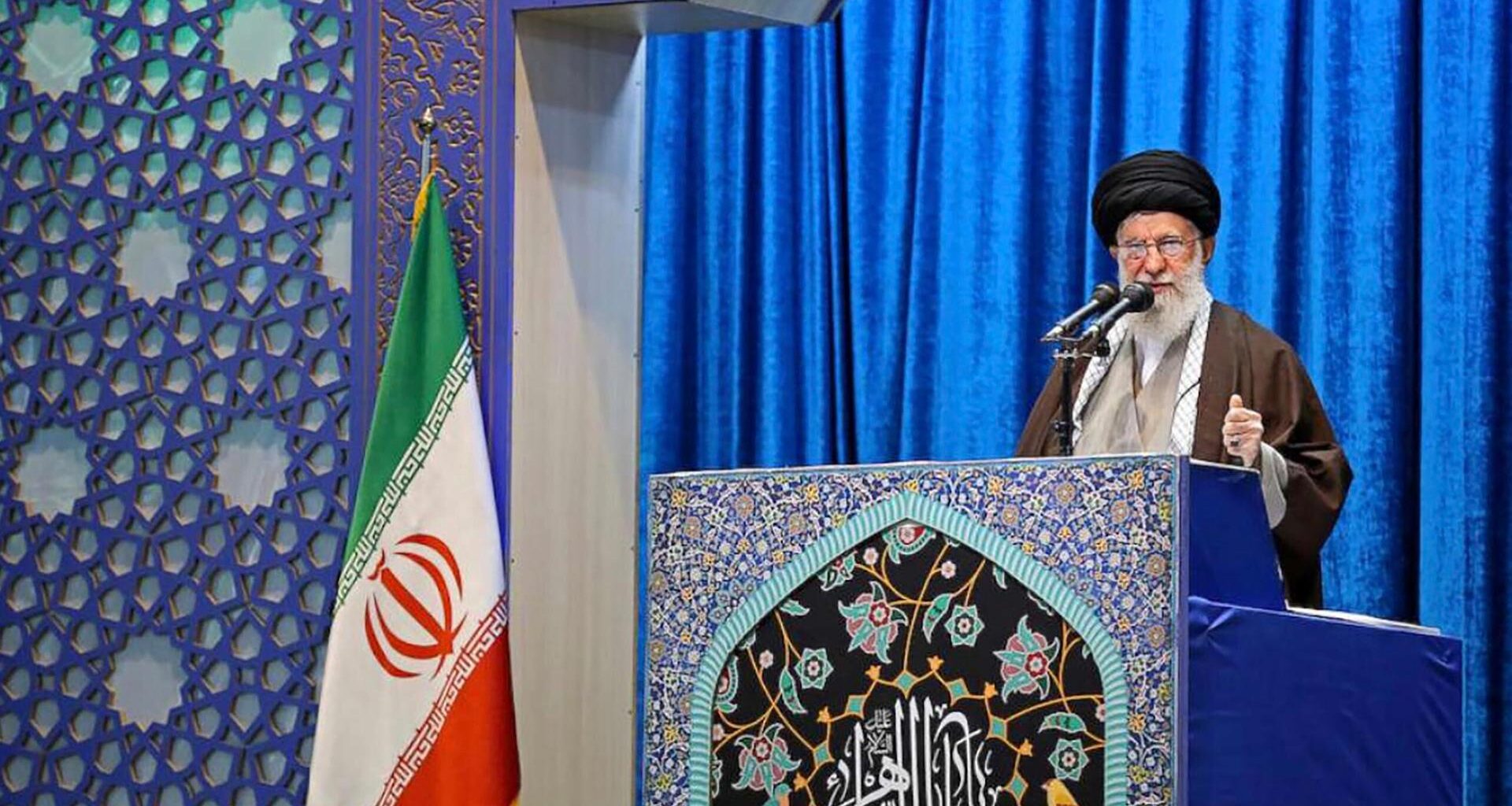 Ayatalloh Ali Khamenei bei Rede vor blauem Samthintergrund neben iranischer Flagge