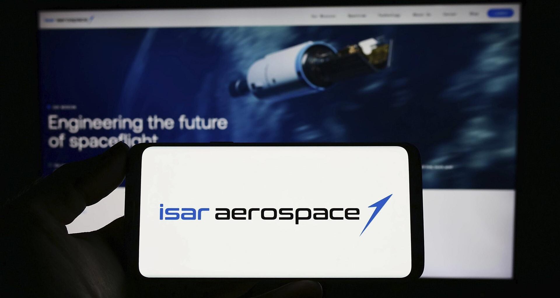 Zu sehen ist das Logo von Isar Aerospace auf einem Smartphone. Auf einem Bildschirm mit der Website des Unternehmens sieht man eine Rakete und den Schriftzug '"Engineering the future of spaceflight".