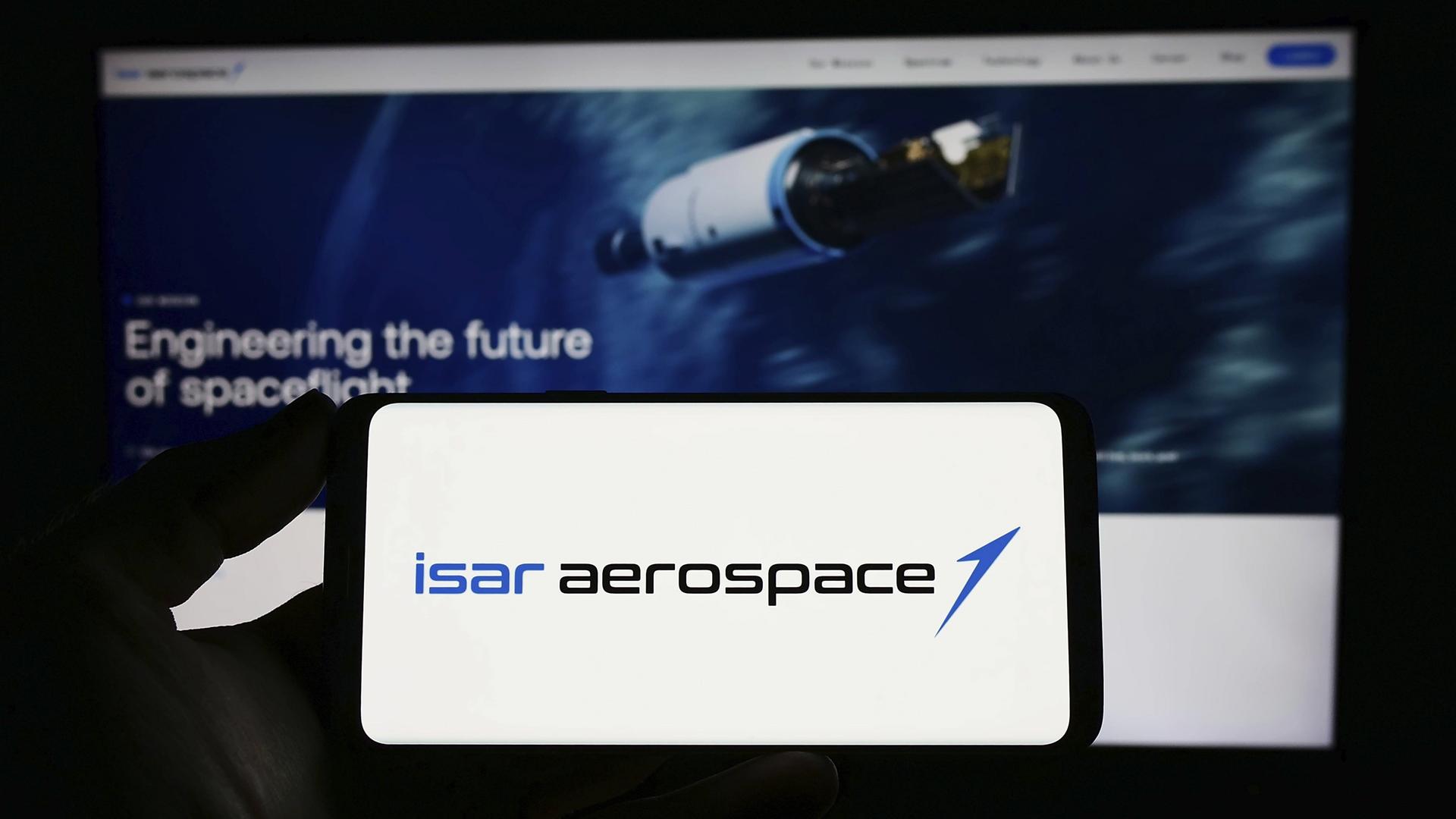 Zu sehen ist das Logo von Isar Aerospace auf einem Smartphone. Auf einem Bildschirm mit der Website des Unternehmens sieht man eine Rakete und den Schriftzug '"Engineering the future of spaceflight".