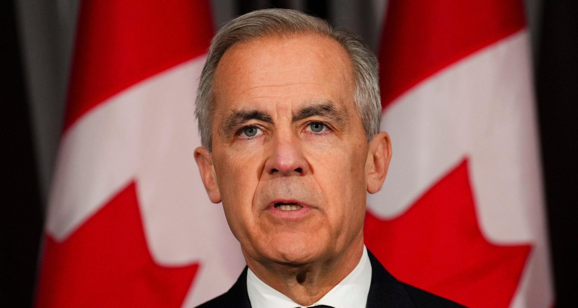 Der kanadische Premierminister Mark Carney steht im dunklen Anzug vor kanadischen Fahnen.