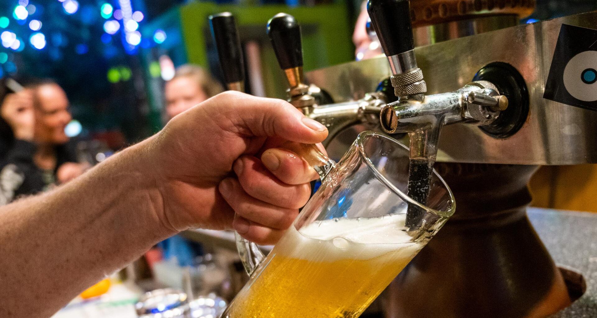 Nahaufnahme: In einer Bar zapft eine Hand Bier in eine Glas. Im HIntergrund unscharf Gäste.