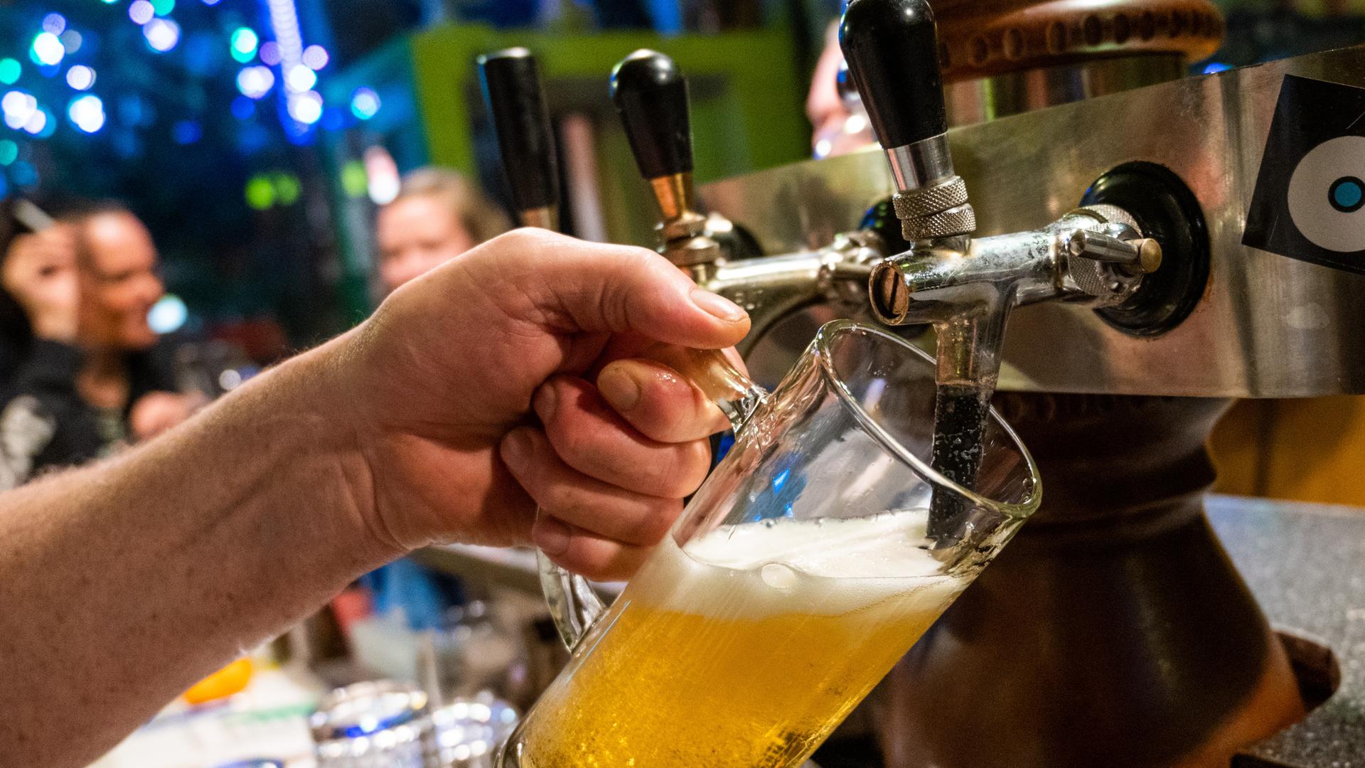 Nahaufnahme: In einer Bar zapft eine Hand Bier in eine Glas. Im HIntergrund unscharf Gäste.