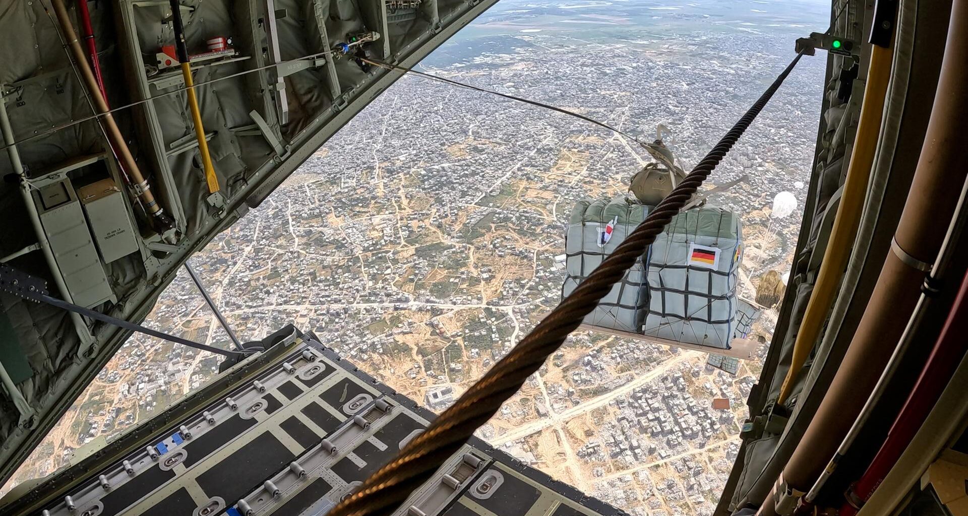 Eine Palette mit Hilfsgütern im freien Fall über dicht besiedeltem Gebiet - aufgenommen durch die Heckklappe eines Transportflugzeugs. Die Reißleine, die den Fallschirm öffnet, ist noch mit dem Flugzeug verbunden.