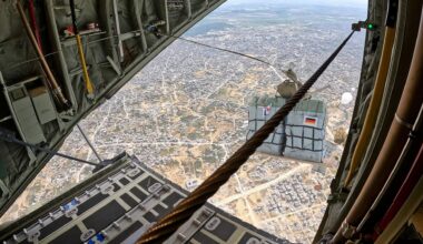Eine Palette mit Hilfsgütern im freien Fall über dicht besiedeltem Gebiet - aufgenommen durch die Heckklappe eines Transportflugzeugs. Die Reißleine, die den Fallschirm öffnet, ist noch mit dem Flugzeug verbunden.