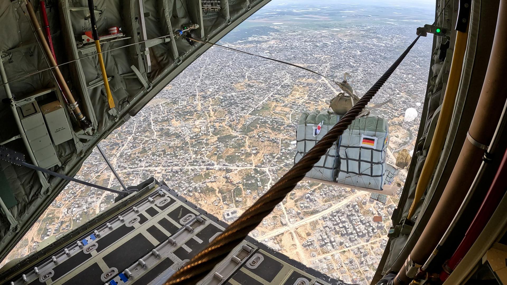 Eine Palette mit Hilfsgütern im freien Fall über dicht besiedeltem Gebiet - aufgenommen durch die Heckklappe eines Transportflugzeugs. Die Reißleine, die den Fallschirm öffnet, ist noch mit dem Flugzeug verbunden.