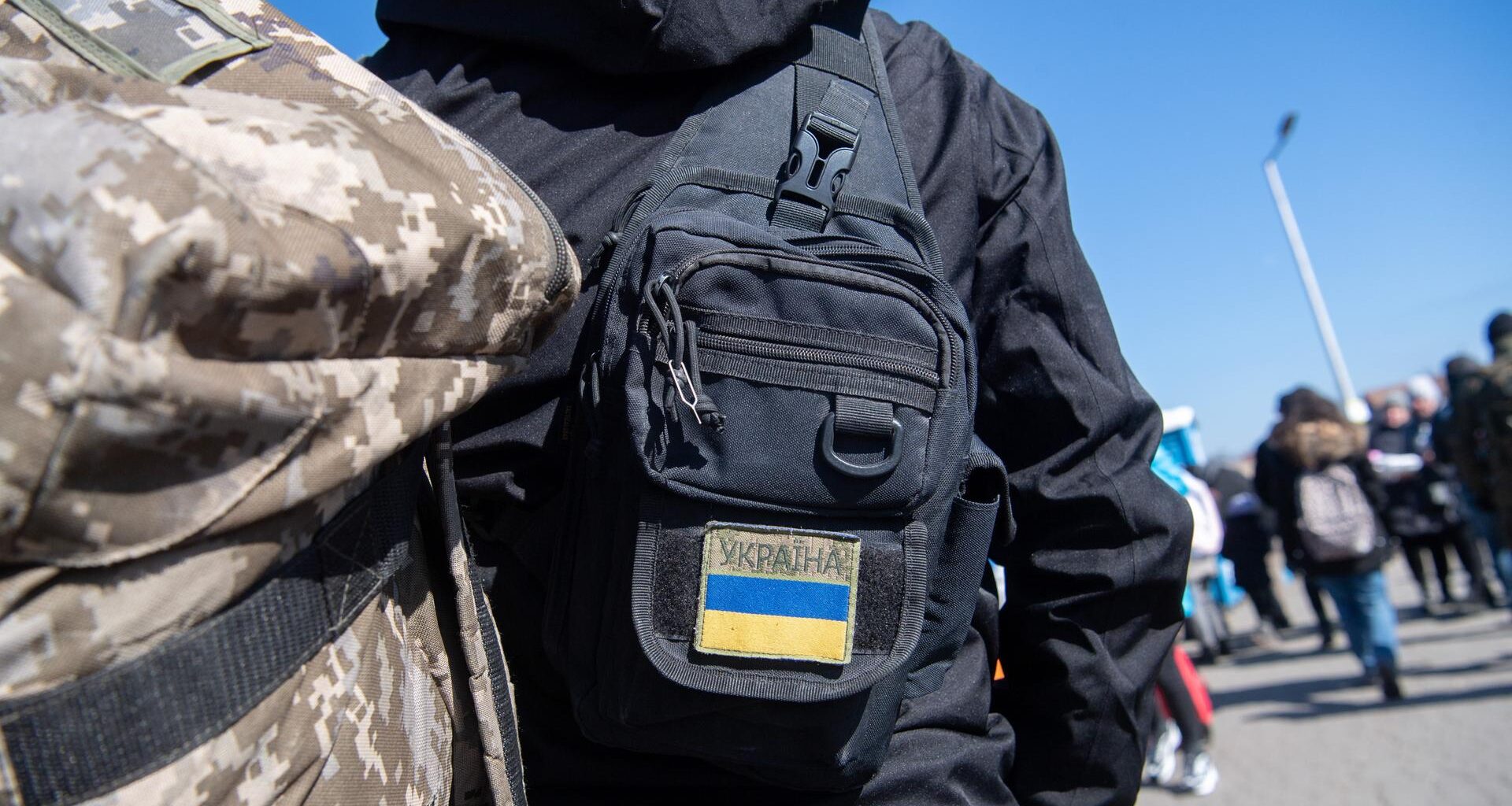 Menschen am polnischen Grenzübergang zur Ukraine, im Vordergrund ein Rücken mit Tarnfleck-Rucksack und einer Tasche mit Ukraine-Flaggenaufnäher