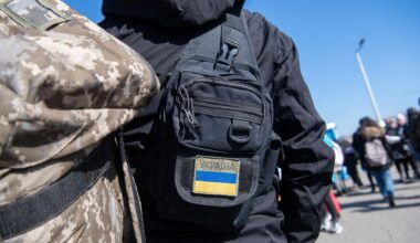 Menschen am polnischen Grenzübergang zur Ukraine, im Vordergrund ein Rücken mit Tarnfleck-Rucksack und einer Tasche mit Ukraine-Flaggenaufnäher