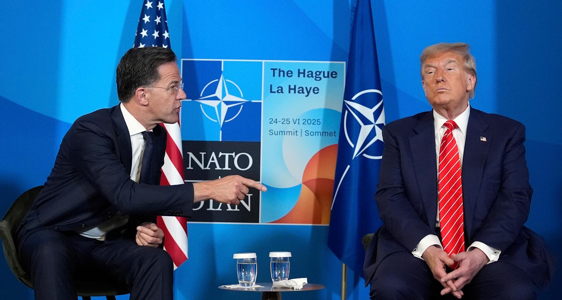 Mark Rutte wendet sich Donald Trump mit ausgstrecktem Zeigefinger zu. Im Hintergrund sieht man die NATO-Flagge.