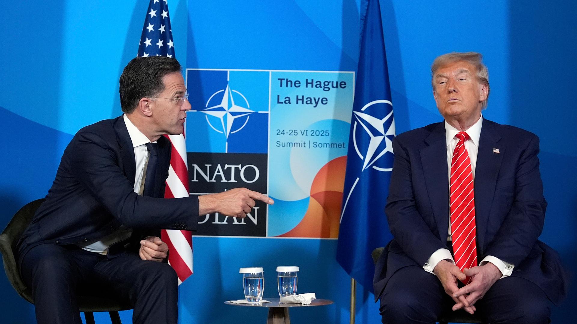 Mark Rutte wendet sich Donald Trump mit ausgstrecktem Zeigefinger zu. Im Hintergrund sieht man die NATO-Flagge.