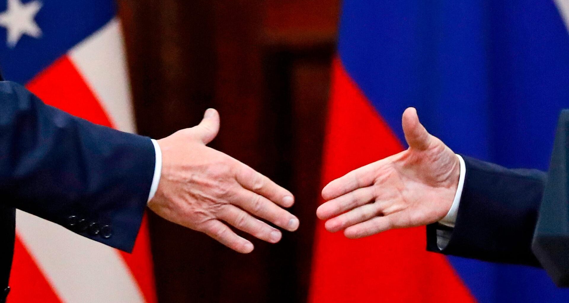 US-Präsident Donald Trump und Russlands Machthaber Wladimir Putin geben sich die Hände: Großaufnahme der Hände, im Hintergrund die russische und US-amerikanische Flagge.