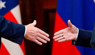 US-Präsident Donald Trump und Russlands Machthaber Wladimir Putin geben sich die Hände: Großaufnahme der Hände, im Hintergrund die russische und US-amerikanische Flagge.
