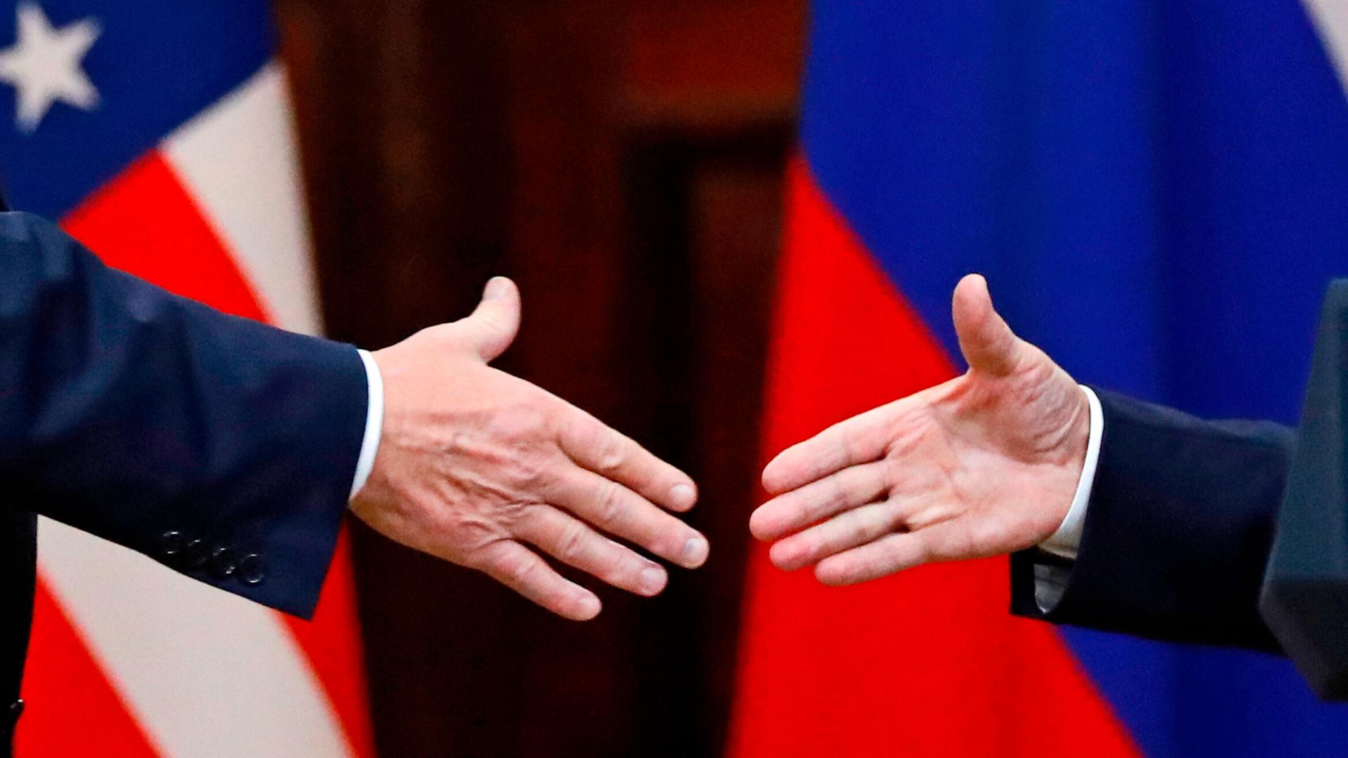 US-Präsident Donald Trump und Russlands Machthaber Wladimir Putin geben sich die Hände: Großaufnahme der Hände, im Hintergrund die russische und US-amerikanische Flagge.