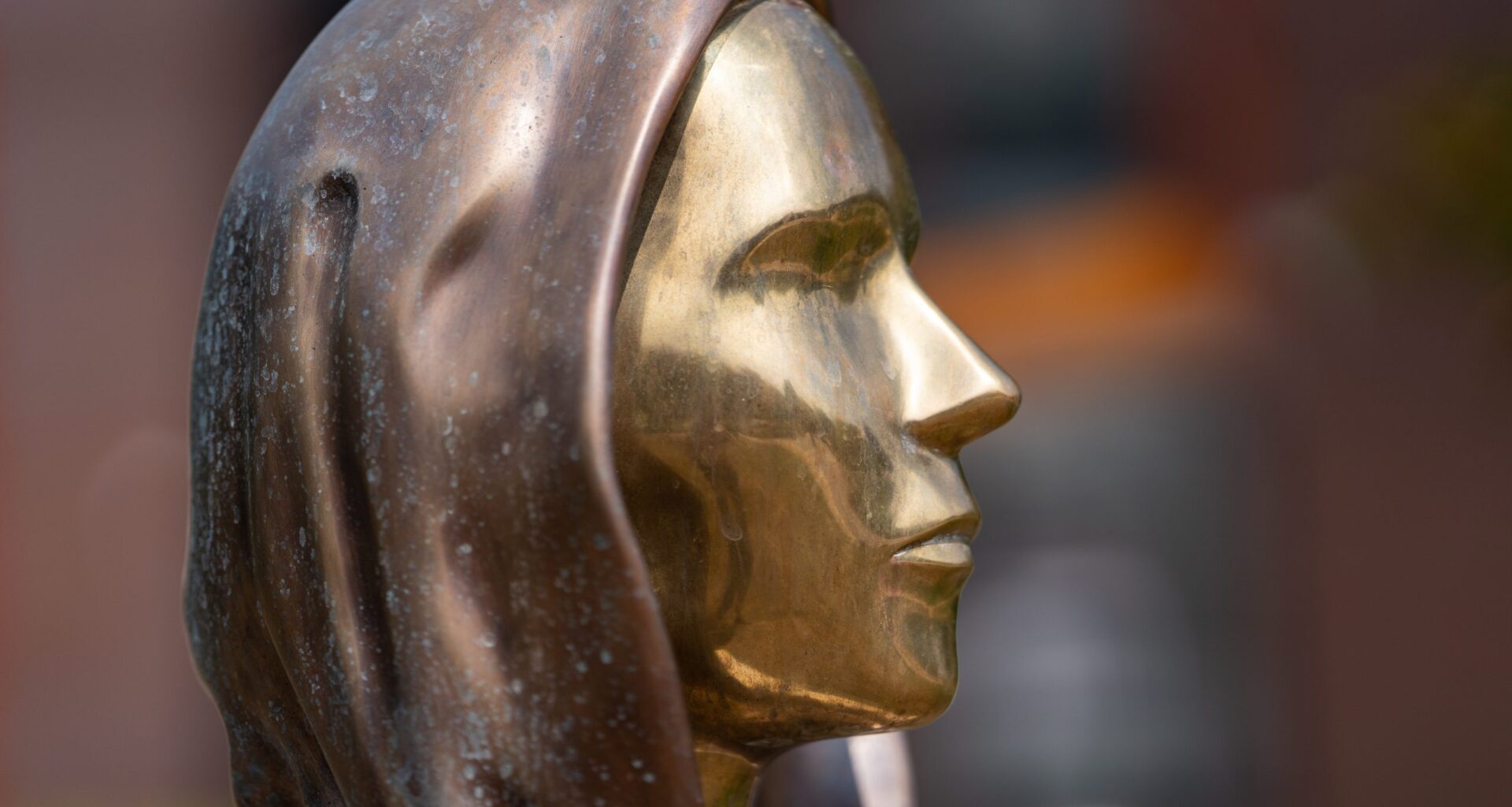 Seitenansicht einer Metallstatue, die Satoshi Nakomoto mit einem menschlichen Gesicht mit Kapuze darstellt, mit einer glatten, reflektierenden goldenen Oberfläche und einer bronzefarbenen Kapuze.